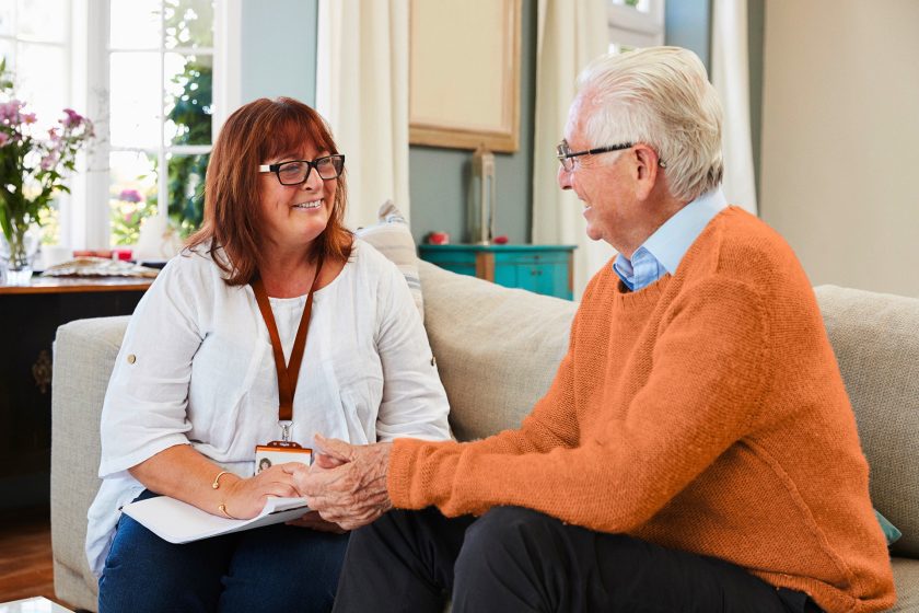 a support worker is assisting an elderly gentleman. They are seated on a couch discussing something.